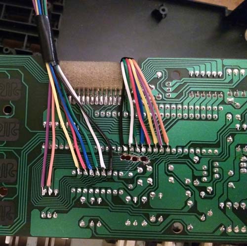 A green printed circuit board in close-up reveals multiple colored wires soldered onto it. The wires, bundled together at the top, fan out to connect with various points on the board. Intricate circuitry paths are visible on this partially disassembled component.