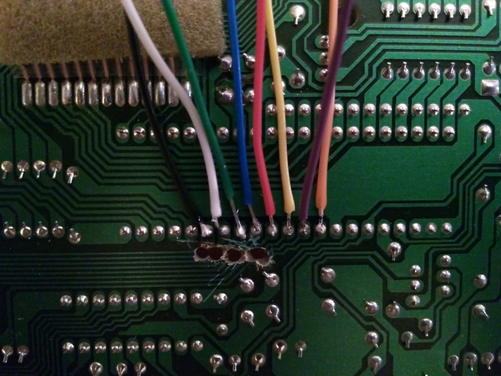 The green circuit board showcases a variety of colorful wires—including white, green, blue, red, yellow, and orange—soldered onto different metal contact points. It features numerous intricate pathways and soldered joints along with a small strip of felt-like material positioned at the top.