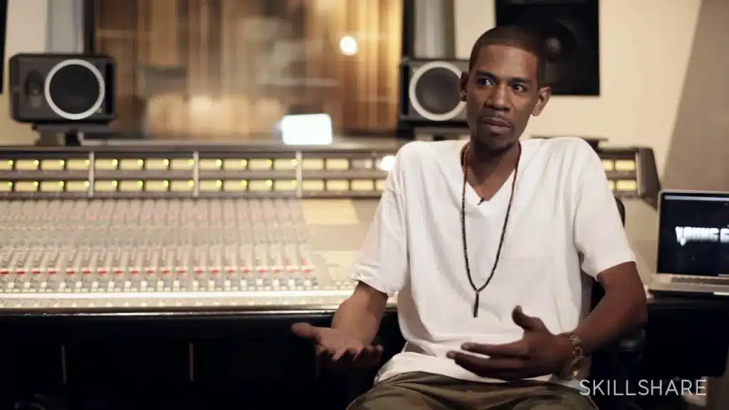 Young Guru in a white T-shirt and necklace sits in front of a large mixing console in a recording studio, gesturing animatedly with their hands as they speak. Nearby, an open laptop rests to the side. The background is filled with speakers and other studio equipment, adding to the professional atmosphere of the space.