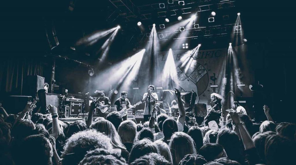 In a black-and-white photo of a live concert, a rock band performs on stage with guitars and microphones. Spotlights shine on the band members, while in the foreground, a large crowd with hands raised enjoys the energetic atmosphere.