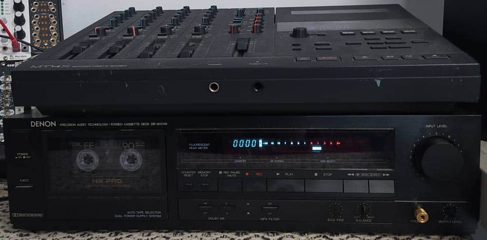 A stack of two audio devices placed one above the other features a multi-channel audio mixer on top, complete with numerous sliders and knobs. Below it sits a vintage Denon cassette deck, equipped with analog meters and various buttons and dials.