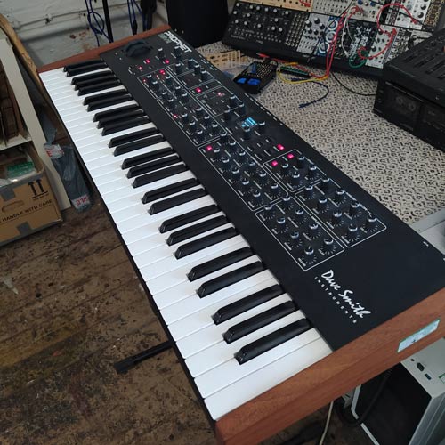 The Prophet Rev 2 synthesizer keyboard, adorned with numerous knobs and buttons, rests on a wooden stand within a busy workspace filled with other electronic gear. Bearing the name "Dave Smith Instruments," the synth features wooden sides along with its traditional white and black keys.