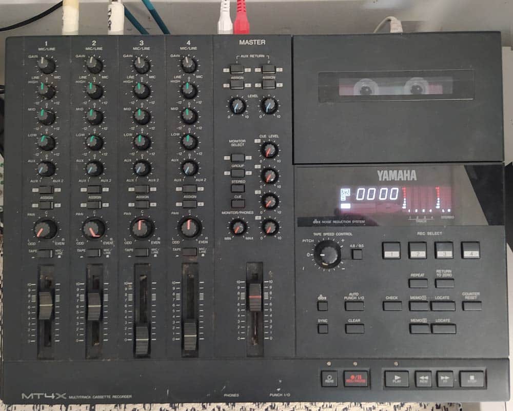 The Yamaha MT4X multitrack cassette recorder displays an array of controls, including various knobs, sliders, and buttons designed for precise adjustments. At the right side, a cassette tape is inserted, while a small digital display indicates numerical information. Connected at the top are multiple cables facilitating its operation.