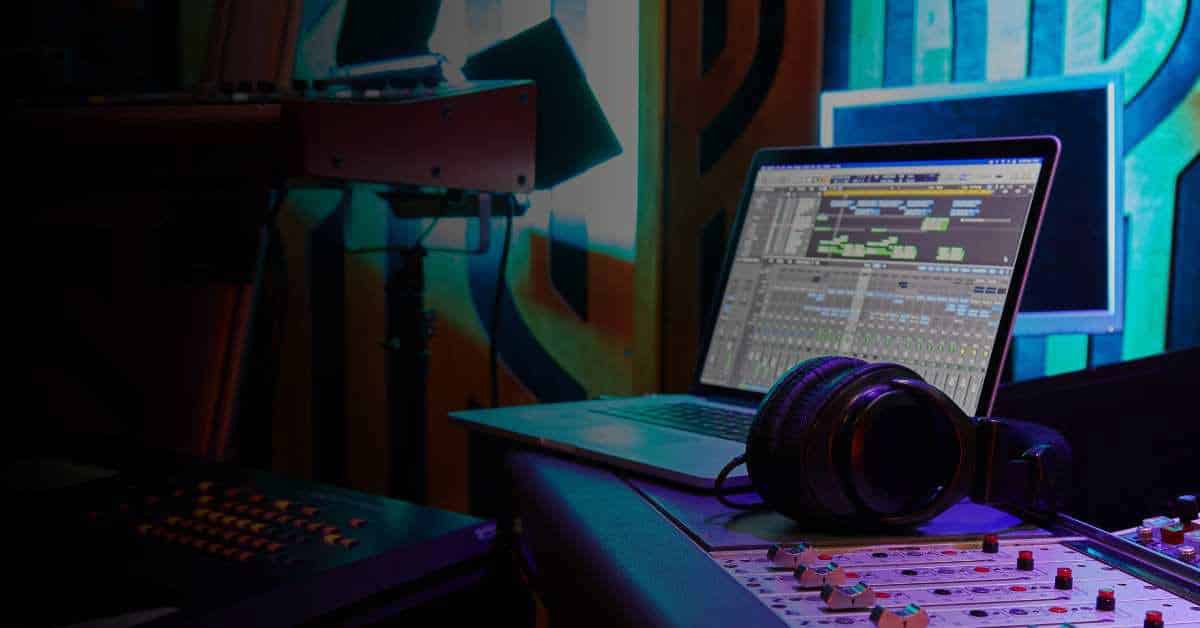 In a dimly lit music studio, a laptop displaying music production software sits on a desk. Nearby, a pair of large headphones and part of a sound mixing console can be seen. Colorful soundproofing panels and other audio equipment are visible in the background.