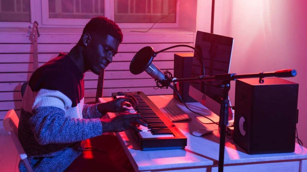 A person is seated at a desk playing a keyboard piano in a music studio. They are wearing a sweater and are surrounded by various pieces of equipment including a microphone, speakers, and a computer. The room is illuminated with a mix of pink and purple lighting.