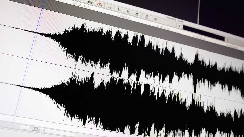 A close-up of a computer screen reveals an audio editing software interface featuring two tracks. Each track presents a waveform with peaks and valleys, illustrating the amplitude of the audio over time. In the background, toolbars and a timeline are displayed.