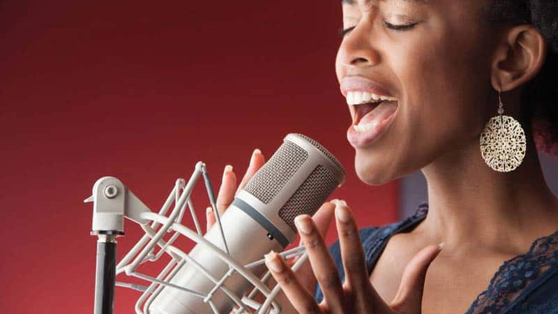 A person with short hair passionately sings into a studio microphone, eyes closed and fully immersed in their performance. Large, circular earrings accentuate their appearance against the solid red background, enhancing the intensity of the scene.